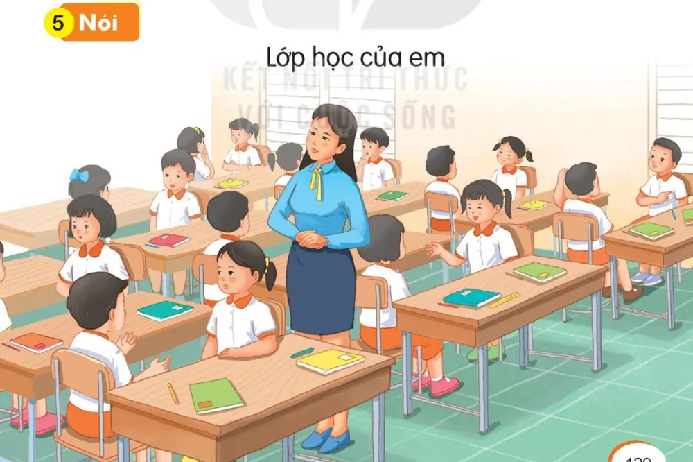 cô giáo đứng giữa lớp, học sinh ngồi ở bàn học.